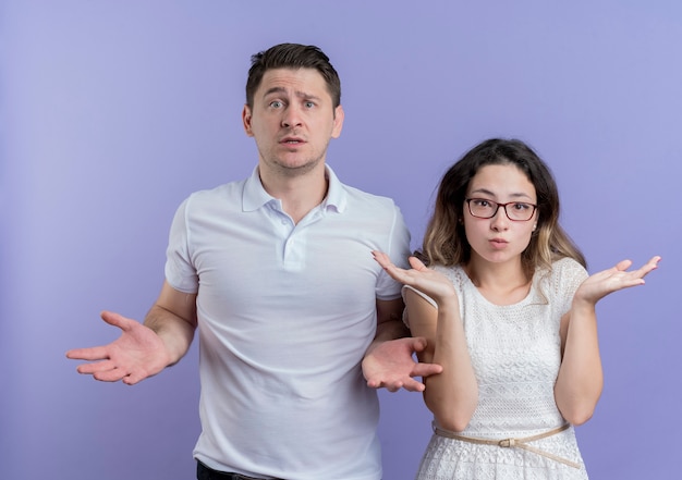 카메라를보고 젊은 부부 남자와 여자는 파란색 벽 위에 서있는 대답이없는 측면에 팔을 확산 혼란