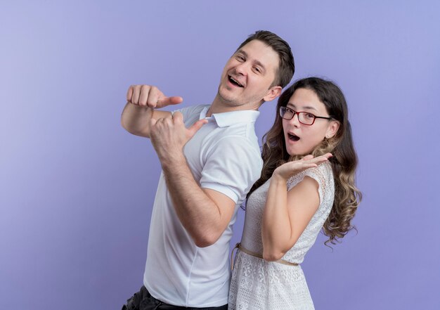 Young couple man and woman looking at camera smiling cheerfully showing thumbs up standing together over blue wall