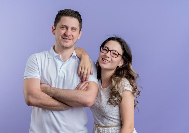 Young couple man and woman looking at camera standing together smiling cheerfully over blue wall