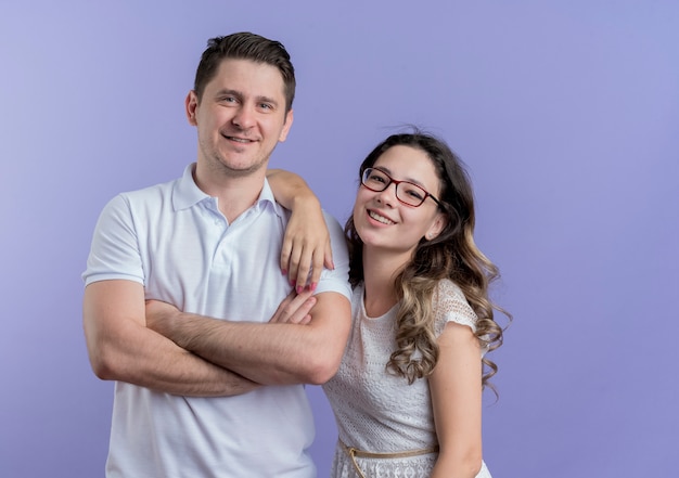 Young couple man and woman looking at camera standing together smiling cheerfully over blue wall