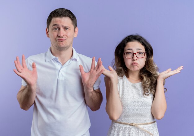 Young couple man and woman looking confused shrugging shoulders standing over blue wall