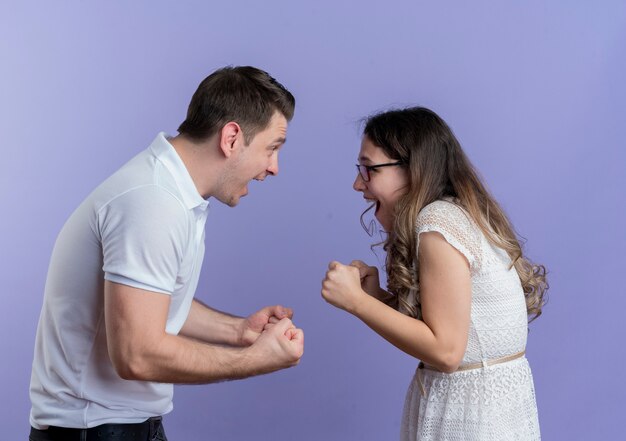 Young couple man and woman looking at each other clenching fists standing over blue wall