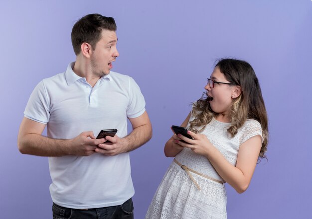 Young couple man and woman looking at each other surprised holding smartphones together over blue
