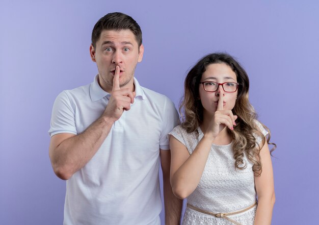 young couple man and woman making silence gesture with fingers on lips over blue