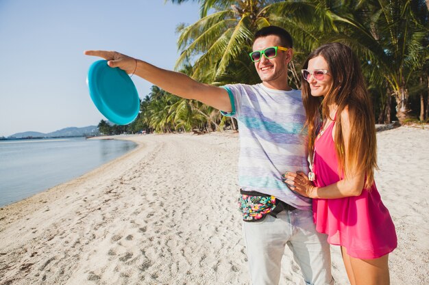 Young couple man and woman playing flying disk on tropical beach, summer vacation, love, romance, happy mood, smiling, having fun, hipster outfit, sunglasses, denim shorts, sunny, positive mood