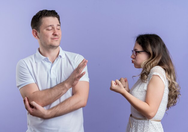 Young couple man and woman quarreling woman with fists looking with angry face at her boyfriend while he showing stop sign standing over blue wall