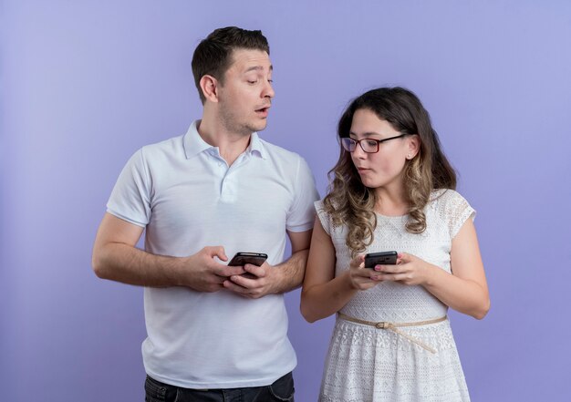 青い壁の上に一緒に立っているスマートフォンを持ってお互いをスパイしている若いカップルの男性と女性