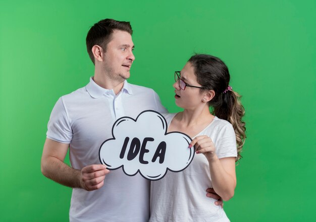 Young couple man and woman standing together smiling happy and positive holding speech bubble sign with word idea looking at each other over green wall