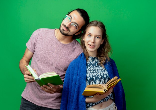 Young couple man and woman with blanket holding books looking at camera smiling standing over green background