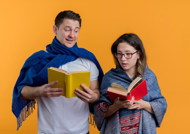 Young couple man and woman with blankets holding books happy and positive smiling standing over orange wall