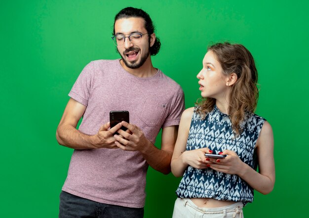 Young couple man and woman with smartphones woman surprised and confused looking at her boyfriend over green background