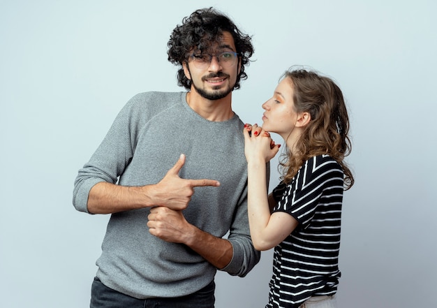 young couple man and woman, woman touching shoulder of her boyfriend while he pointing with finger to her standing over white wall