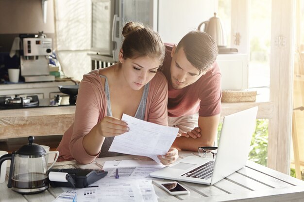 Young couple managing finances, reviewing their bank accounts using laptop computer and calculator at modern kitchen. Woman and man doing paperwork together