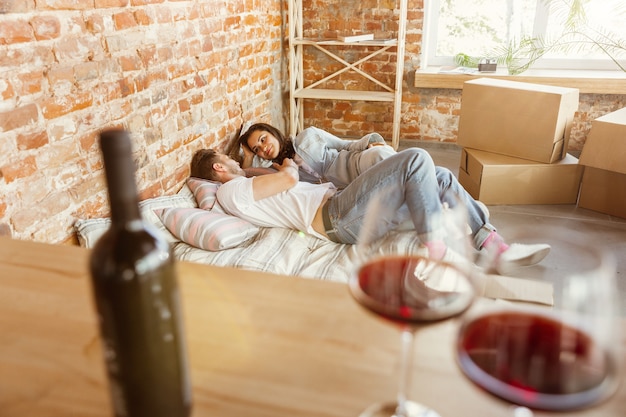 Young couple moved to a new house or apartment. Drinking red wine, caddling and relaxing after cleaning and unpacking. Look happy and confident. Family, moving, relations, first home concept.