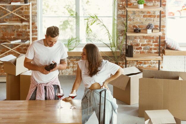 Young couple moved to a new house or apartment. Drinking red wine, smiling and relaxing after cleaning and unpacking. Look happy and confident. Family, moving, relations, first home concept.