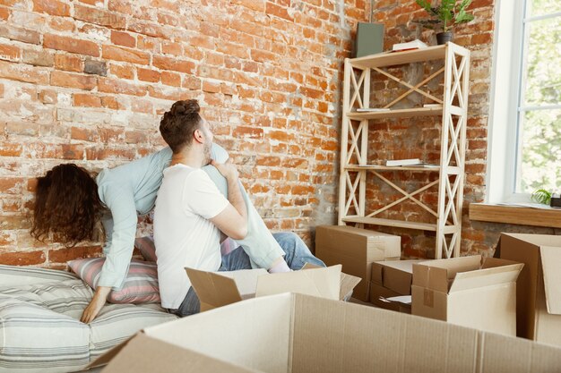 若いカップルは新しい家やアパートに引っ越しました。段ボール箱を一緒に開梱し、引っ越しの日を楽しんでください。幸せで、夢のようで、自信を持って見えます。家族、引っ越し、関係、最初の家のコンセプト。