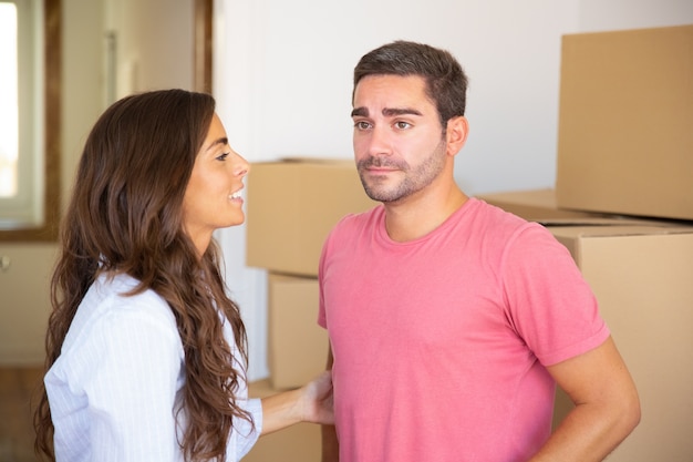 Young couple moving into new apartment, standing among carton boxes and discussing unpacking