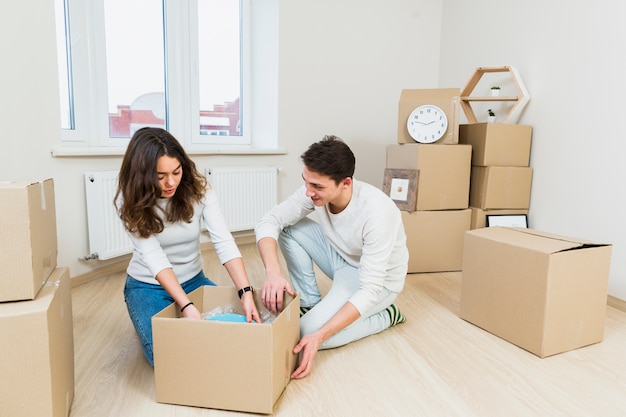 Young couple moving to new place sitting near cardboard box unpacking dishes