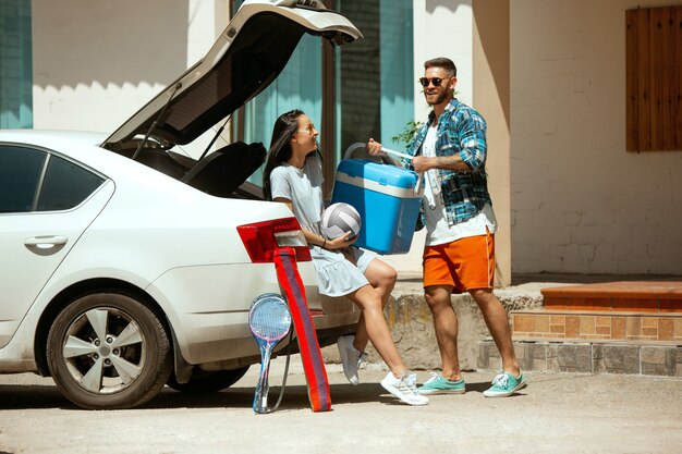 晴れた日に車で休暇旅行の準備をしている若いカップル。スポーツ用品を積み上げる女性と男性。海、川沿い、または海に行く準備ができています。関係の概念、夏、週末。