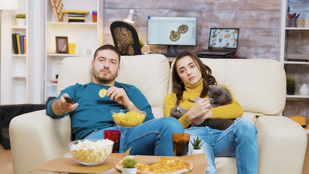 젊은 부부는 TV를 시청하는 고양이와 함께 소파에서 휴식을 취합니다. 칩을 먹는 남자.
