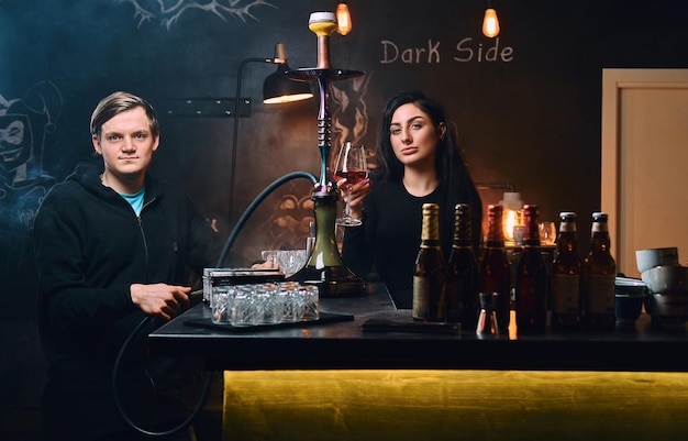 Young couple relaxing at nightclub or bar. Handsome guy in hoodie smokes a hookah and seductive brunette woman drinks wine.