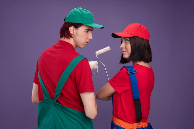 Young couple serious guy confident girl in construction worker uniform and cap standing in behind view holding paint roller looking at each other isolated on purple wall