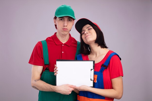 Молодая пара серьезный парень и довольная девушка в форме и кепке строителя держит и показывает парень с закрытыми глазами с закрытыми глазами