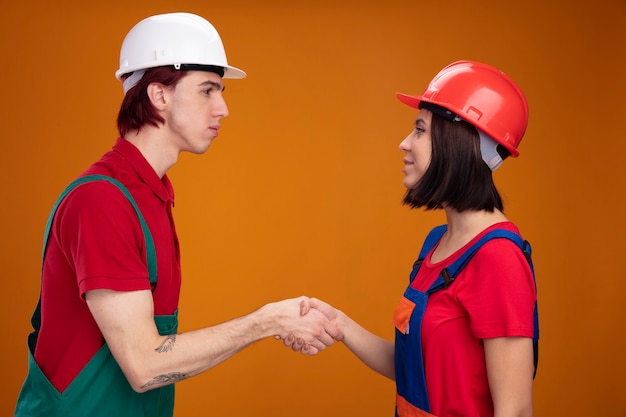 Young couple serious guy pleased girl in construction worker uniform and safety helmet standing in profile view looking at each other greeting isolated on orange wall