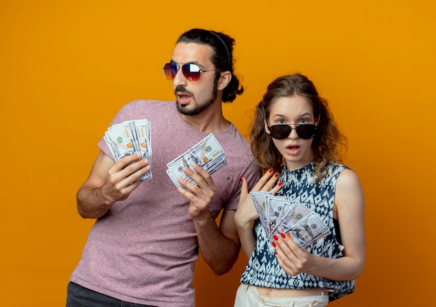 Бесплатное фото Молодая пара показывает наличные деньги, стоящие над оранжевой стеной
