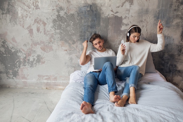 Молодая пара, сидящая на кровати дома в повседневной одежде, занятый мужчина, работающий внештатным сотрудником на ноутбуке, женщина, слушающая музыку в наушниках, проводящая время вместе