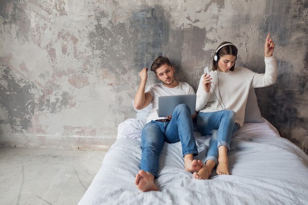 Young couple sitting on bed at home in casual outfit, busy man working freelance on laptop, woman listening to music on headphones, spending time together