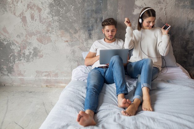 Young couple sitting on bed at home in casual outfit, busy man working freelance on laptop, woman listening to music on headphones, spending time together