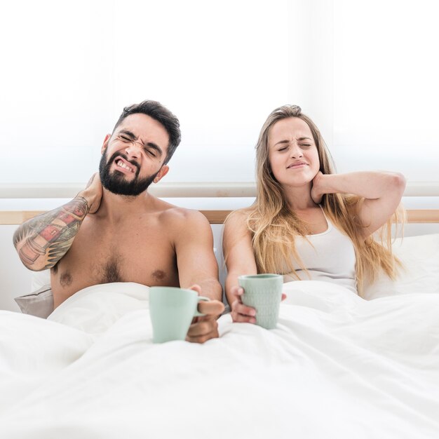 Young couple sitting on bed with cup of coffee suffering from neck pain