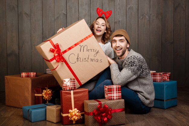 Young couple sitting among christmas gift boxes over wooden wall