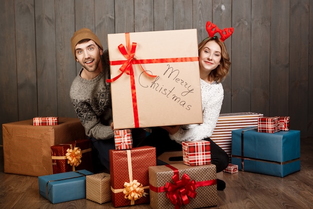 Young couple sitting among christmas gift boxes over wooden wall