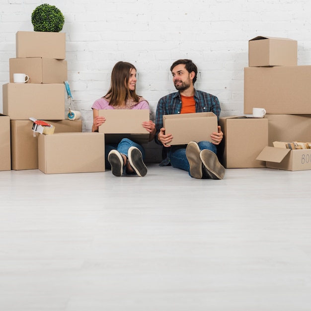 Free photo young couple sitting on floor holding cardboard boxes in hand looking to each other