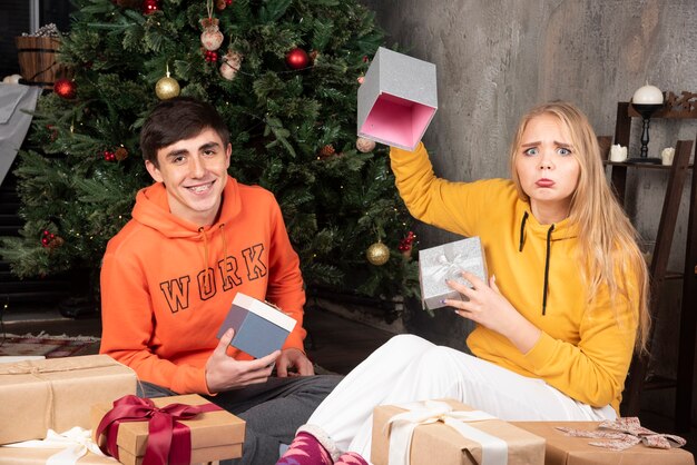 젊은 부부는 바닥에 앉아서 크리스마스 트리 근처에서 선물을 엽니다.
