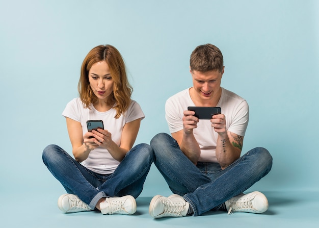 Young couple sitting on floor watching video on cellphone against blue backdrop