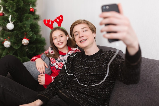 이어폰을 끼고 집에서 소파에 앉아 셀카를 찍고 배경에 크리스마스 트리와 함께 시간을 보내는 젊은 부부
