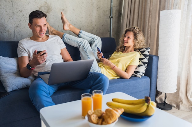 Молодая пара, сидя на диване у себя дома с помощью смартфонов