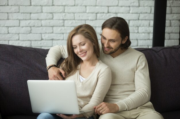 Молодая пара, сидя на диване, обнимая с помощью ноутбука на дому