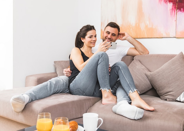 Young couple sitting on sofa watching video on mobile phone at home