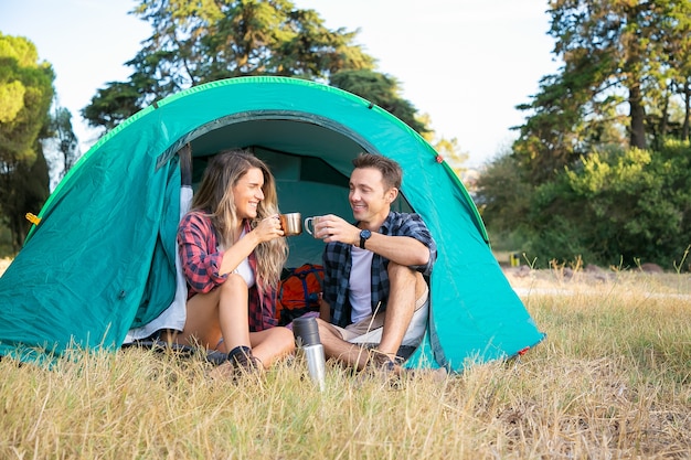 Young couple sitting in tent, talking and drinking tea from thermos. Happy hikers relaxing on lawn, camping, smiling and enjoying nature on weekends. Tourism, adventure and summer vacation concept