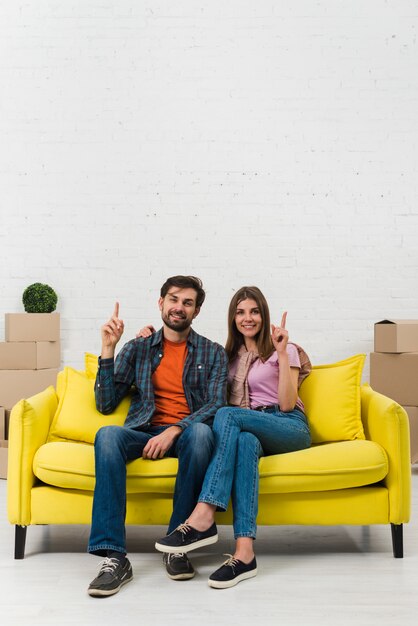 Young couple sitting on yellow sofa pointing their fingers upward and looking to camera