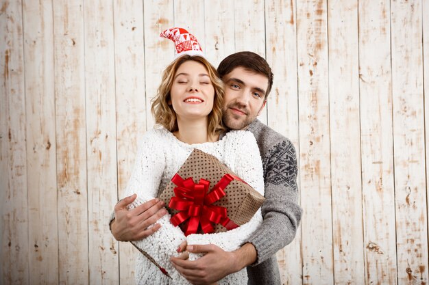 Young couple smiling embracing holding christmas gift over wooden wall