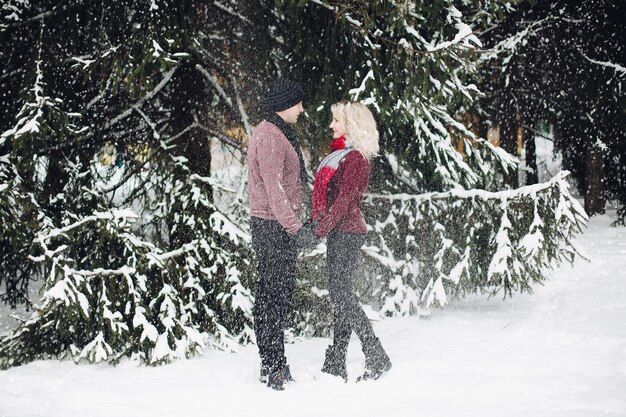 Young couple standing and looking each other in snowing forest. Blonde wife and her husband wearing red sweaters. Woman with volumed hair touching her significant other. Concept of tendernness.