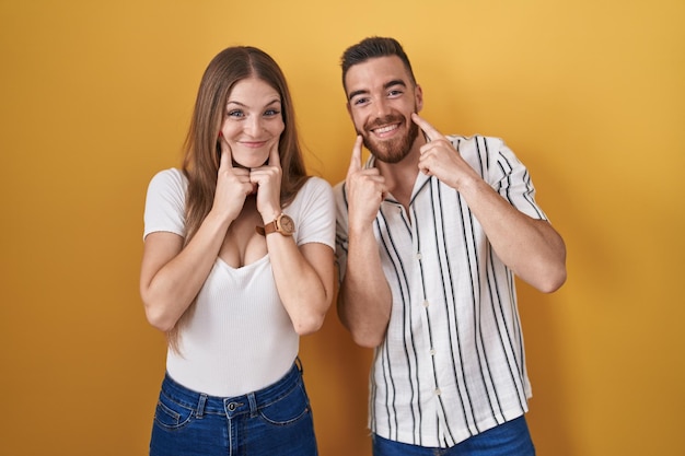 Young couple standing over yellow background smiling with open mouth, fingers pointing and forcing cheerful smile