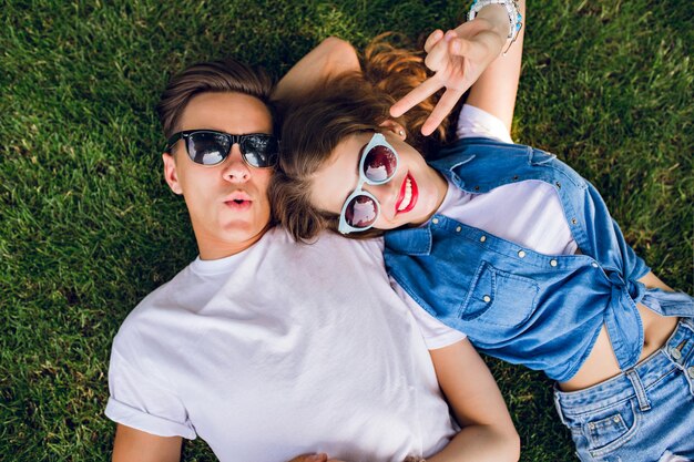 Young couple in sunglasses is lying on grass in park. Girl with long curly hair is lying on shoulder of handsome guy in white T-shirt.  They aping to camera. View from above.