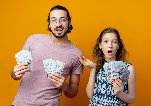 若いカップルはオレンジ色の壁の上に立っている現金を示して驚いた