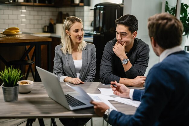 Young couple talking about blueprints while having a meeting with investment agent and using a computer